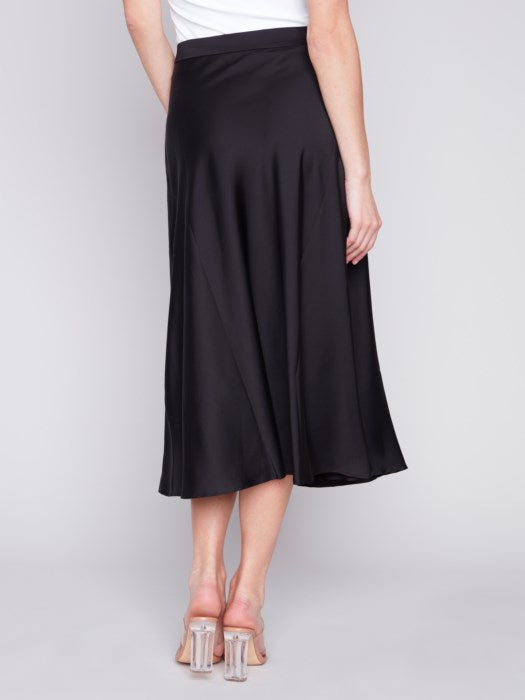 Jupe satin noir pour femme, taille élastique, 97% polyester, 3% spandex. Vue rapprochée  de la jupe portée avec des talons transparents.